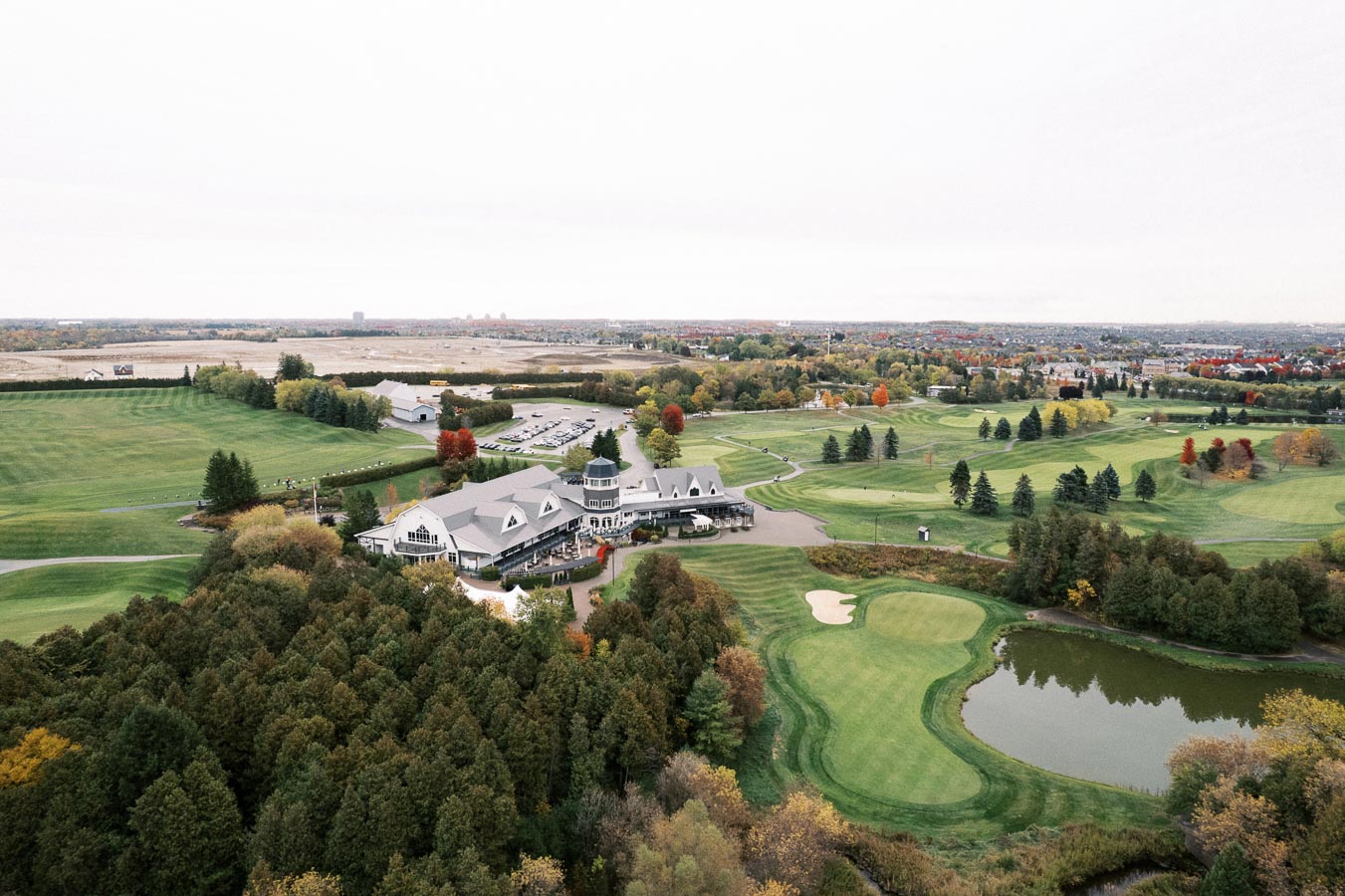Aerial view of a picturesque golf course surrounded by lush greenery and autumnal trees, featuring a large clubhouse, neatly manicured fairways, sand traps, and a reflecting pond.