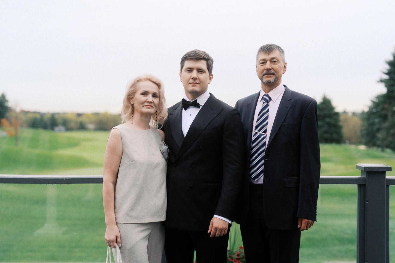 A well-dressed family standing together outdoors with a scenic golf course background, including a woman in a light dress, a man in a tuxedo, and another man in a suit and tie, all smiling.