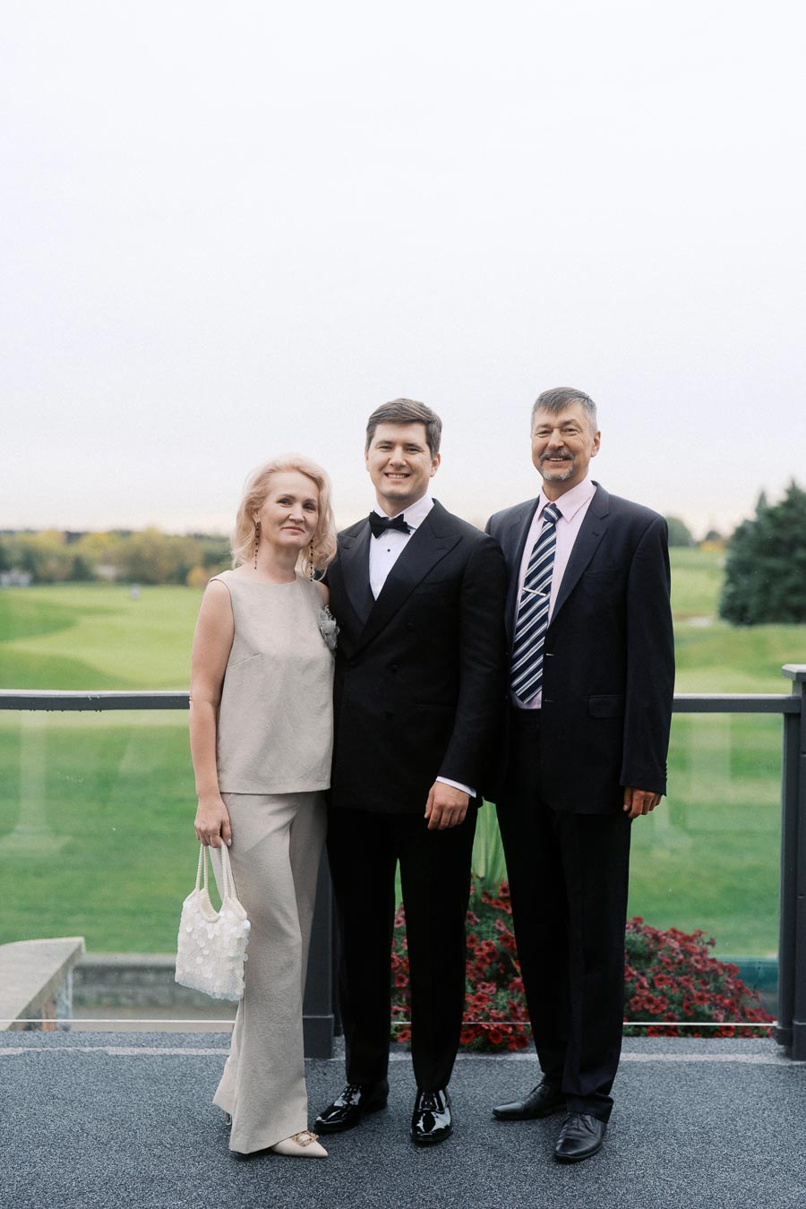 A person in a tuxedo stands between two older adults dressed in formal attire, posing on a balcony overlooking a scenic green landscape with trees in the background.