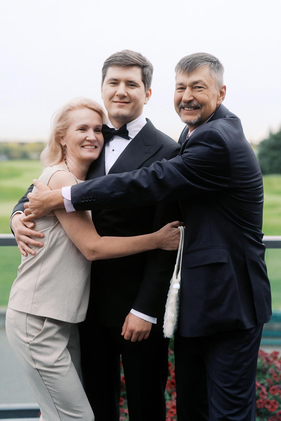 A well-dressed family joyfully embraces outdoors, with a smiling young man in a tuxedo between an older woman and an older man, symbolizing family togetherness.