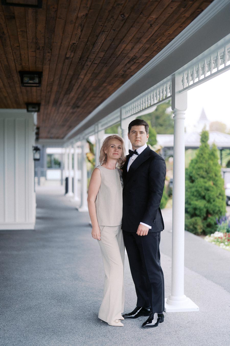 Elegant couple posing under a wooden ceiling with white columns, featuring a woman in a light-colored outfit and a man in a formal black suit, in an outdoor setting with greenery.