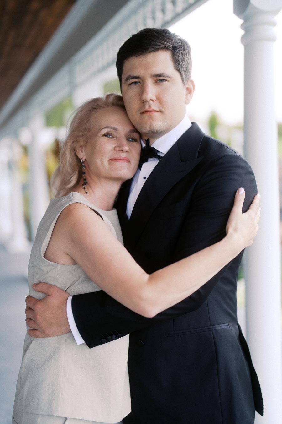 Mother and son embracing on a porch, with the son wearing a black tuxedo and the mother in a beige dress.