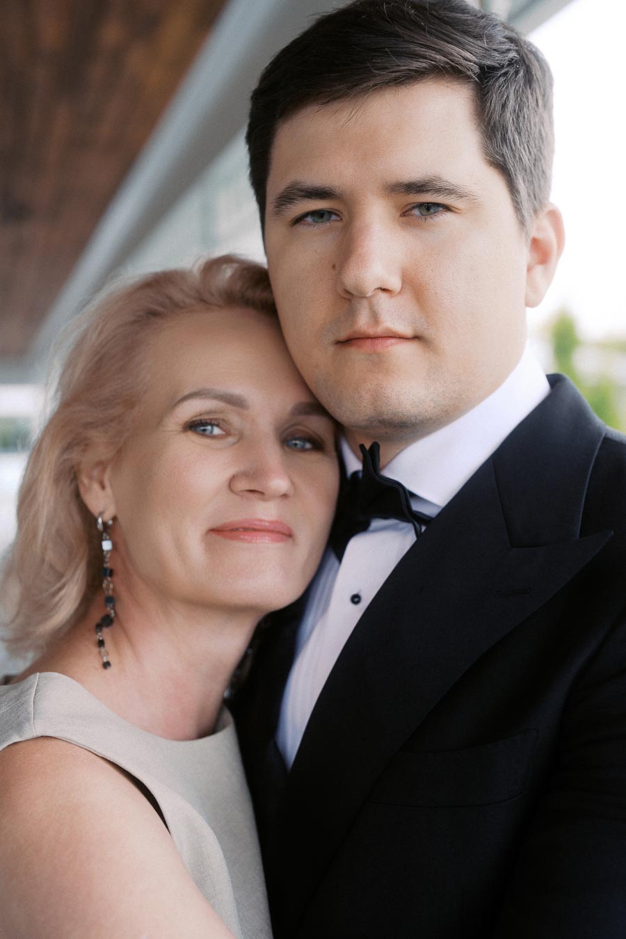 A couple in formal attire, with the woman embracing the man as they pose together on a covered outdoor walkway. The man wears a tuxedo, and the woman is in a sleeveless dress, both looking content and elegant.