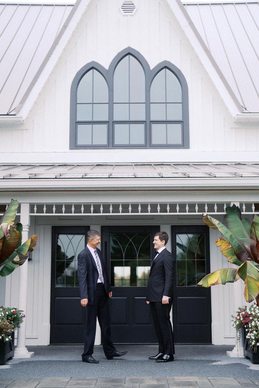 Two men in suits standing and talking in front of an elegant white building with decorative windows and greenery.
