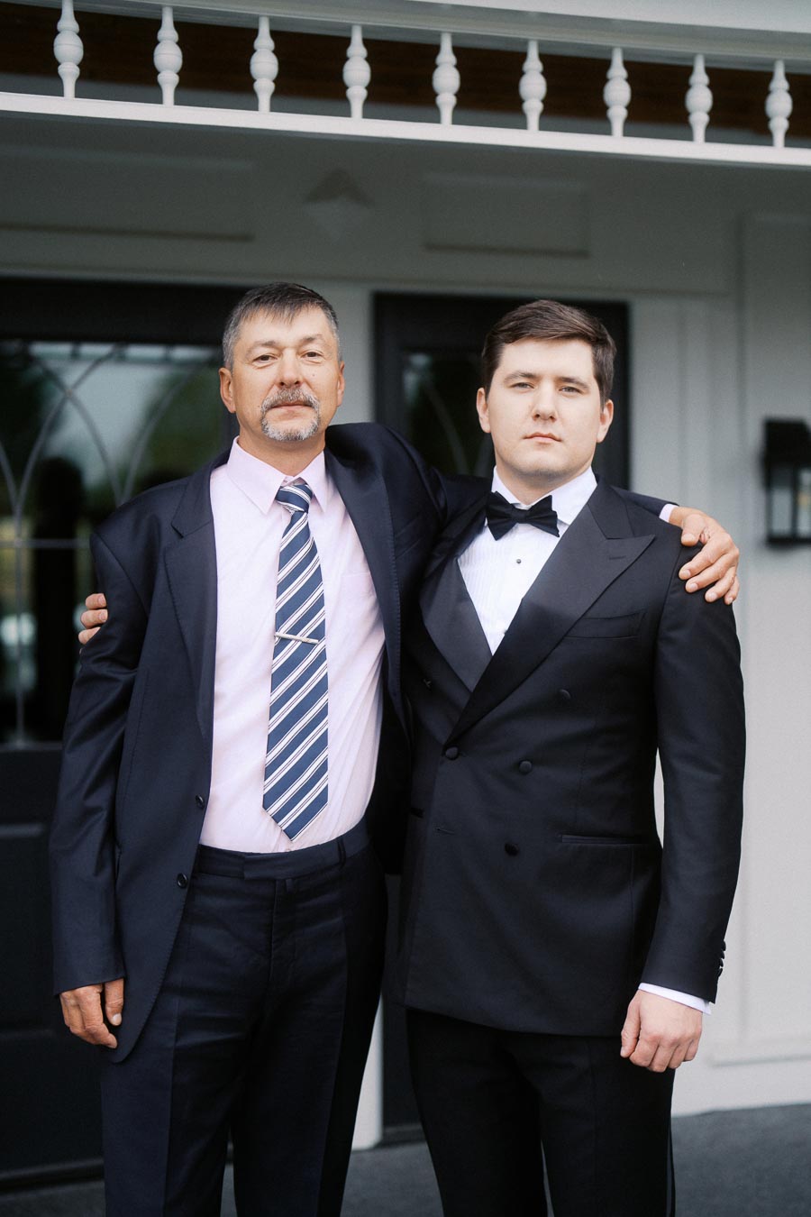 Two men in formal attire, posing together outdoors in front of a building with decorative railing. One man is wearing a pink shirt and striped tie, while the other is in a black tuxedo. They appear to be attending a formal event.