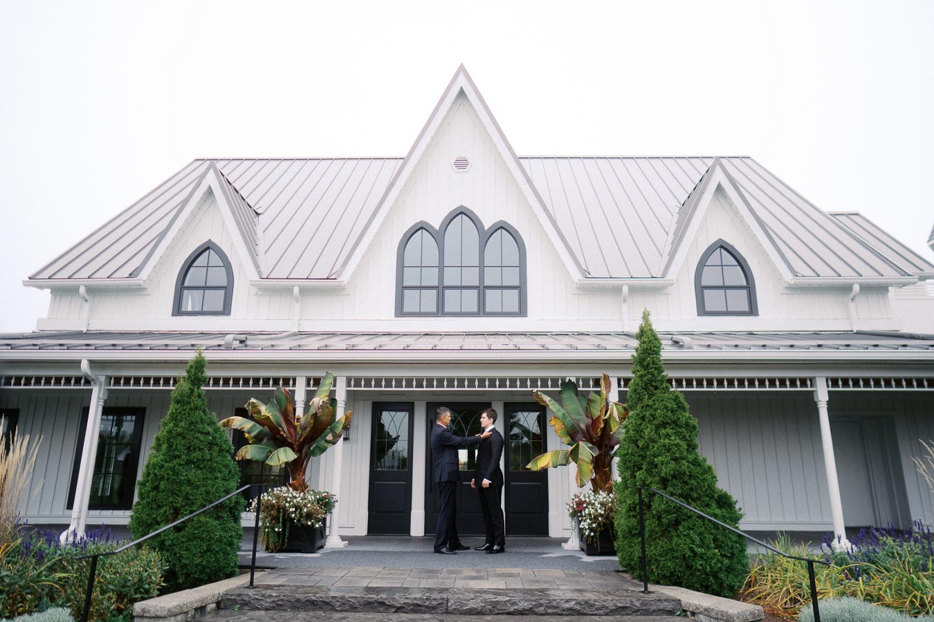 Elegant country house with Gothic-style windows, featuring two men shaking hands on the front porch surrounded by lush greenery.