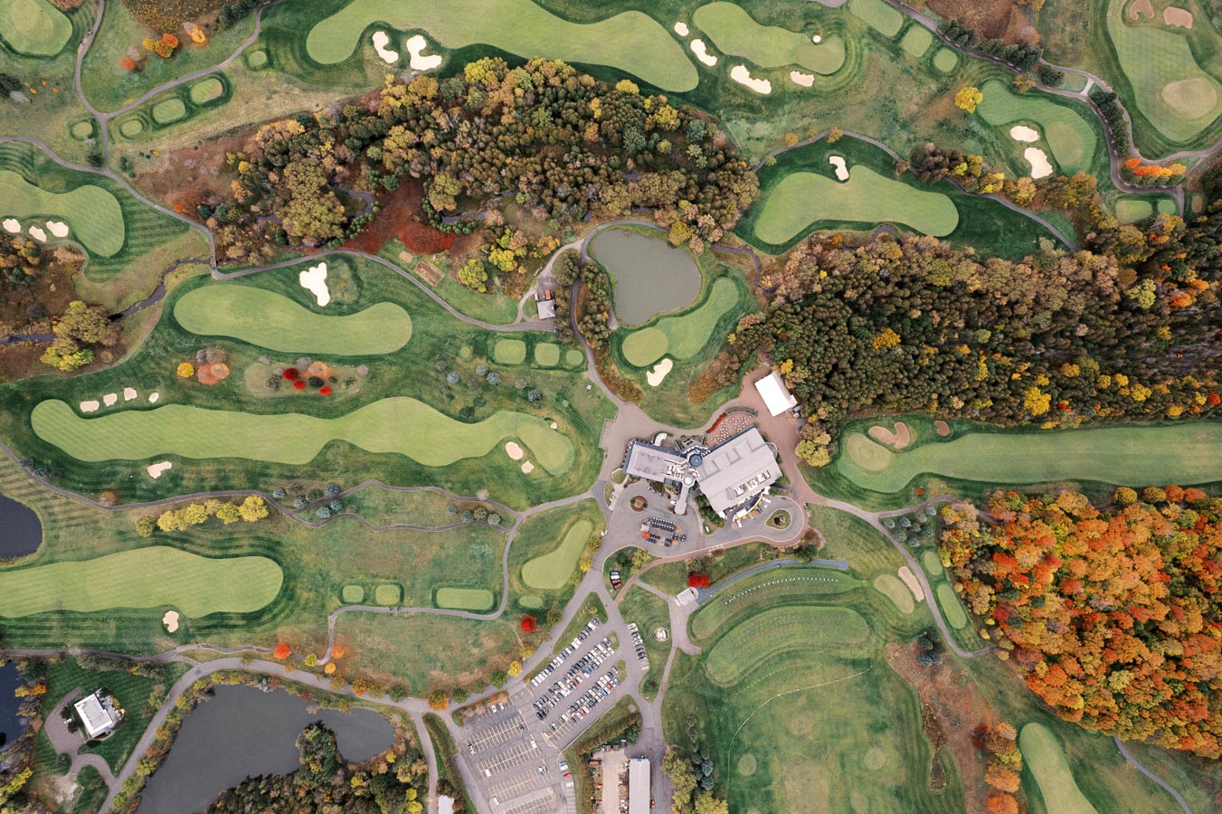 Aerial view of a golf course showcasing lush green fairways, sand bunkers, and water hazards surrounded by autumn foliage, highlighting a clubhouse and parking area at the center.