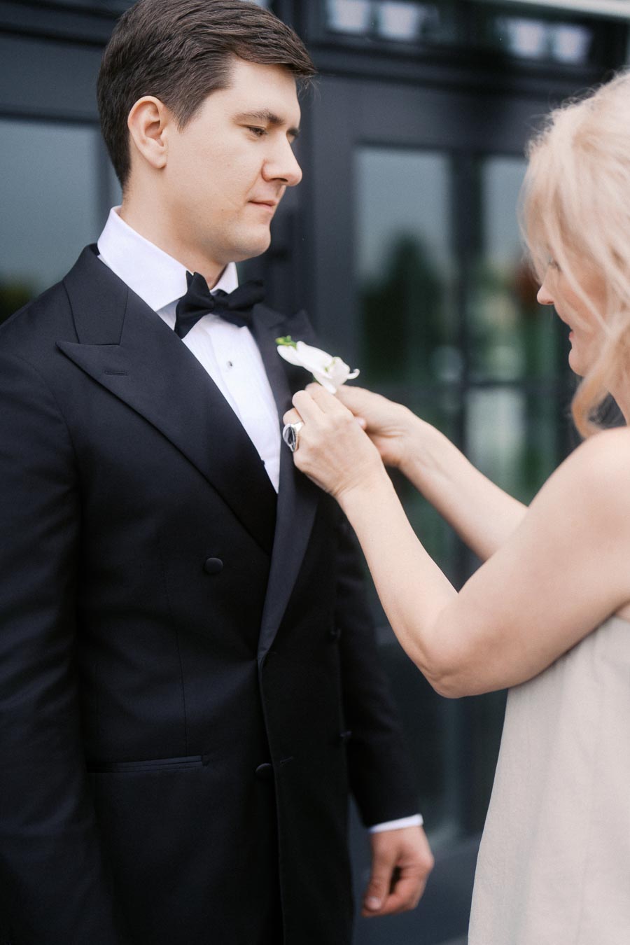 A well-dressed man in a black tuxedo has a boutonniere pinned to his lapel by a woman, in preparation for a formal event.