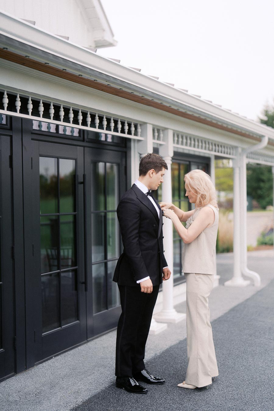 Elegant outdoor wedding preparation scene, featuring a woman adjusting a man's boutonniere on his suit jacket, standing in front of a stylish building with large windows and white detailing.