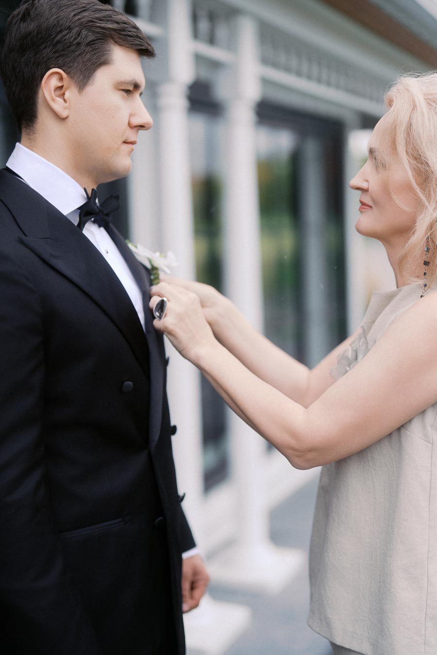 A well-dressed man in a black tuxedo standing outside, while a woman helps adjust his boutonniere.