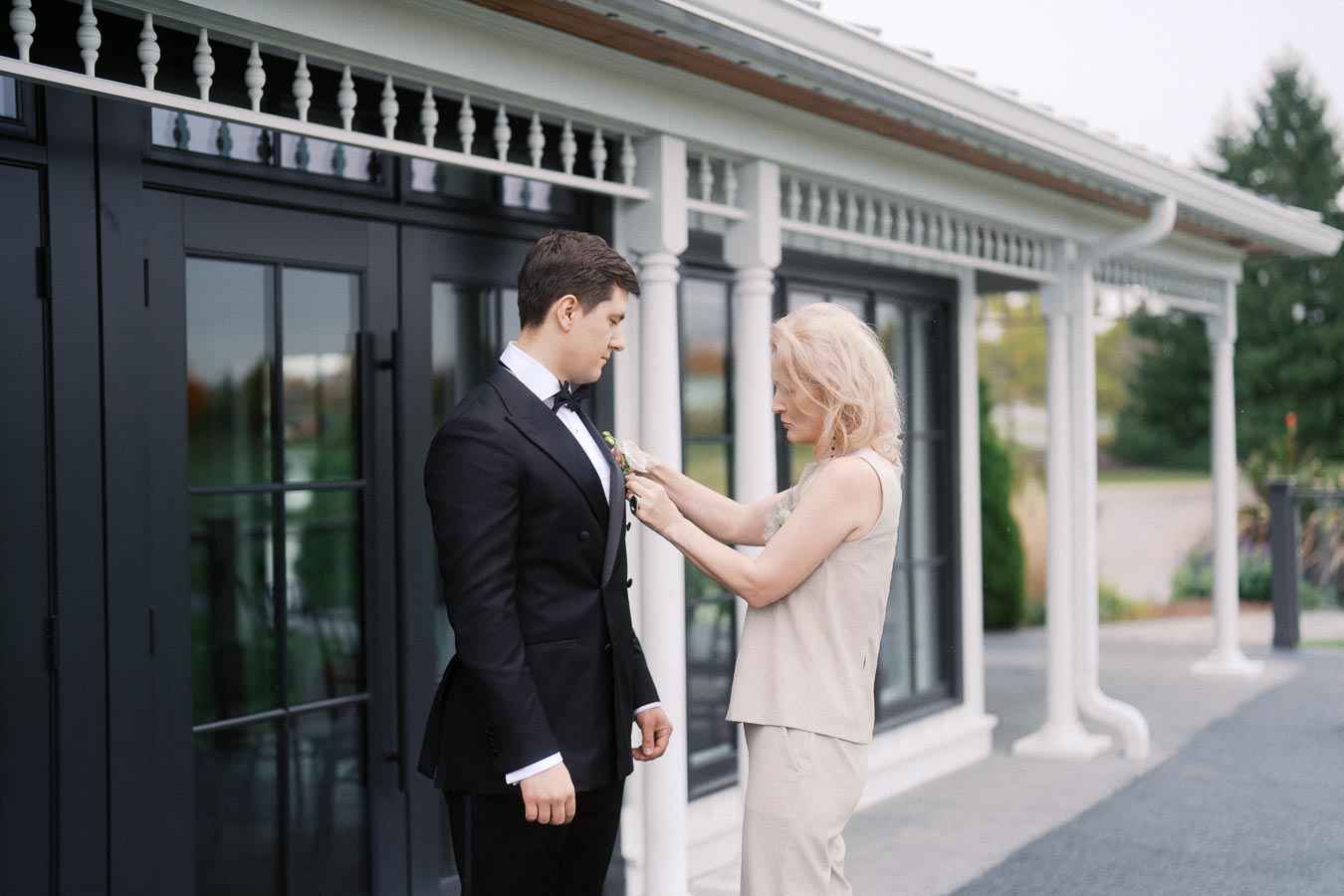 A woman adjusts a boutonniere on a man dressed in a black tuxedo, standing outdoors in front of a modern building with glass and black-framed doors, on what appears to be a formal occasion.