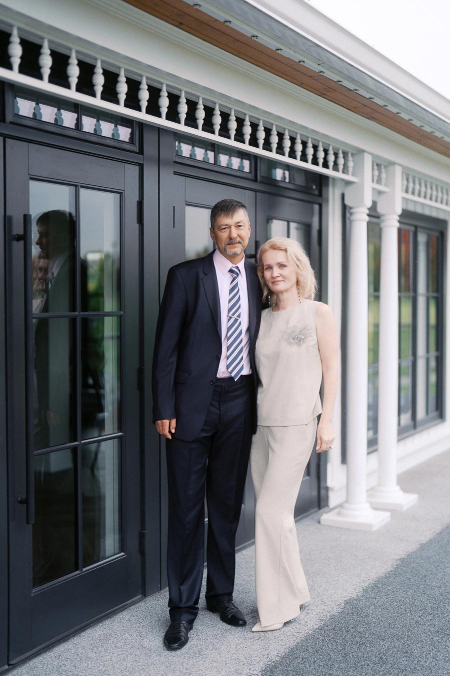 Elegant couple posing together outside a modern building with large glass doors, man in a suit and woman in a beige outfit, showcasing formal attire and companionship.