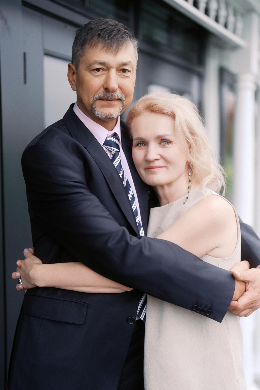 A mature couple embracing lovingly in an elegant setting, with the man wearing a black suit and the woman in a sleeveless dress, conveying warmth and affection.