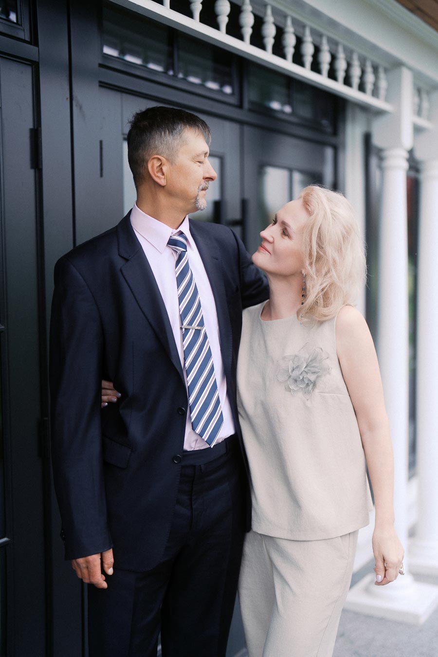 A couple dressed in elegant attire, with the man wearing a dark suit and the woman in a light-colored dress, standing affectionately close outdoors against a stylish building facade.