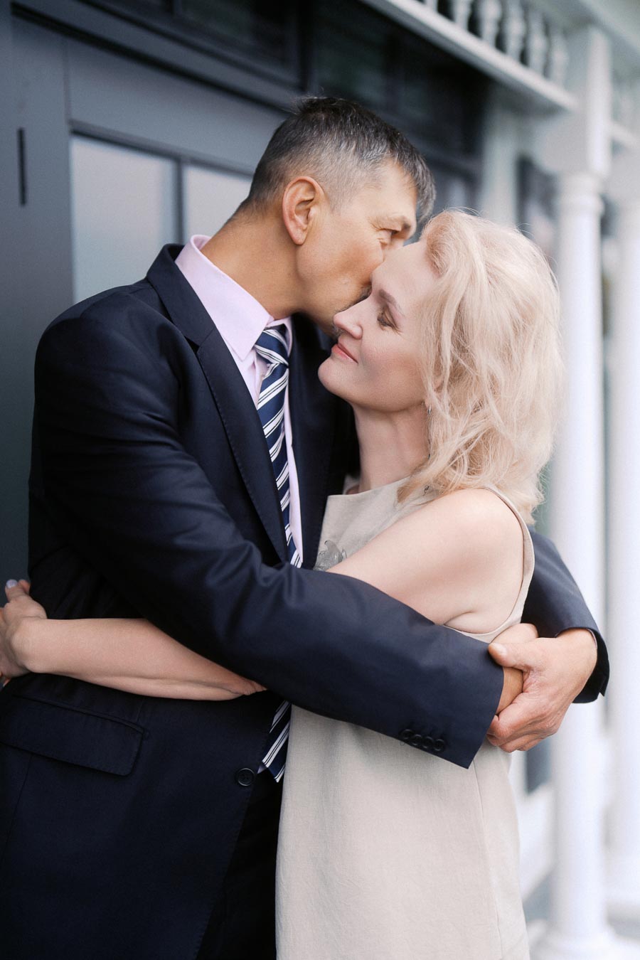 A couple embracing lovingly outdoors, with the man in a suit gently kissing the woman's forehead, conveying affection and intimacy.