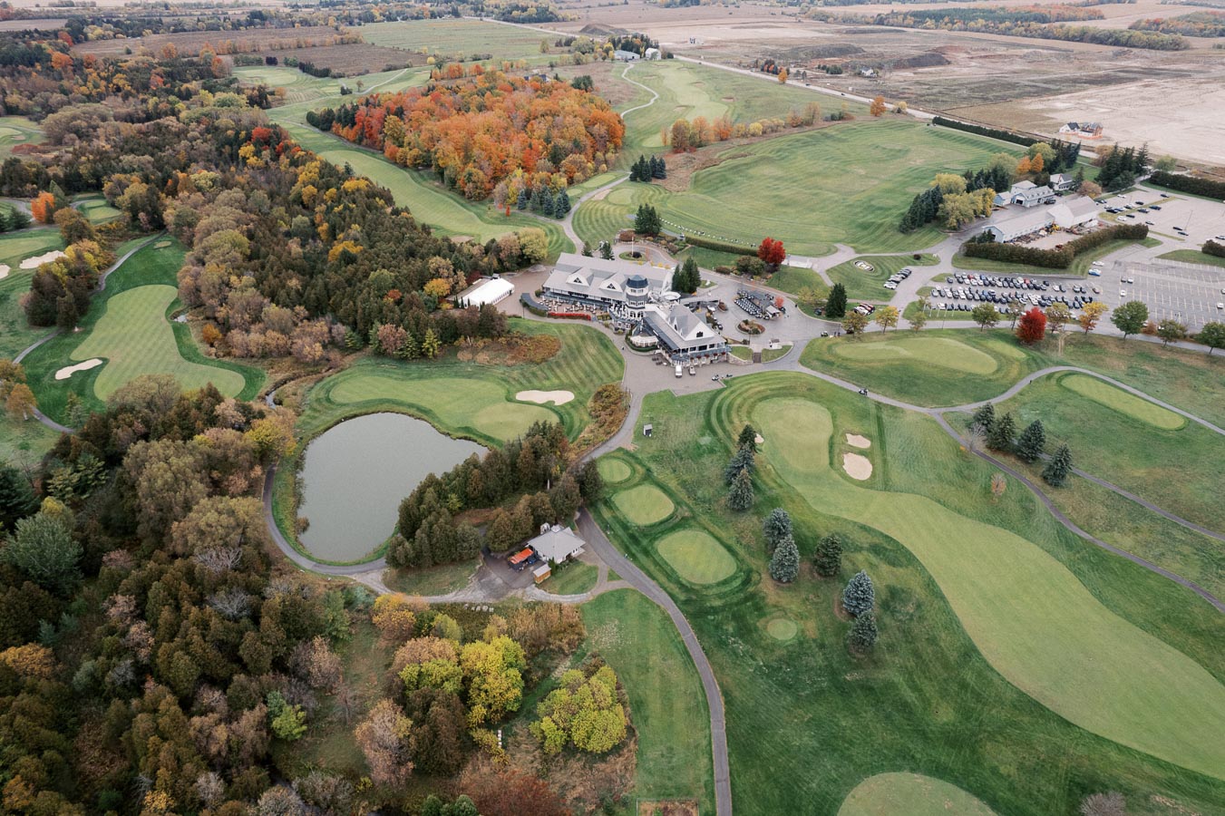 Aerial view of a picturesque golf course surrounded by lush trees in fall colors, featuring well-manicured greens, sand bunkers, and a central clubhouse with adjacent parking, set in a scenic rural landscape.