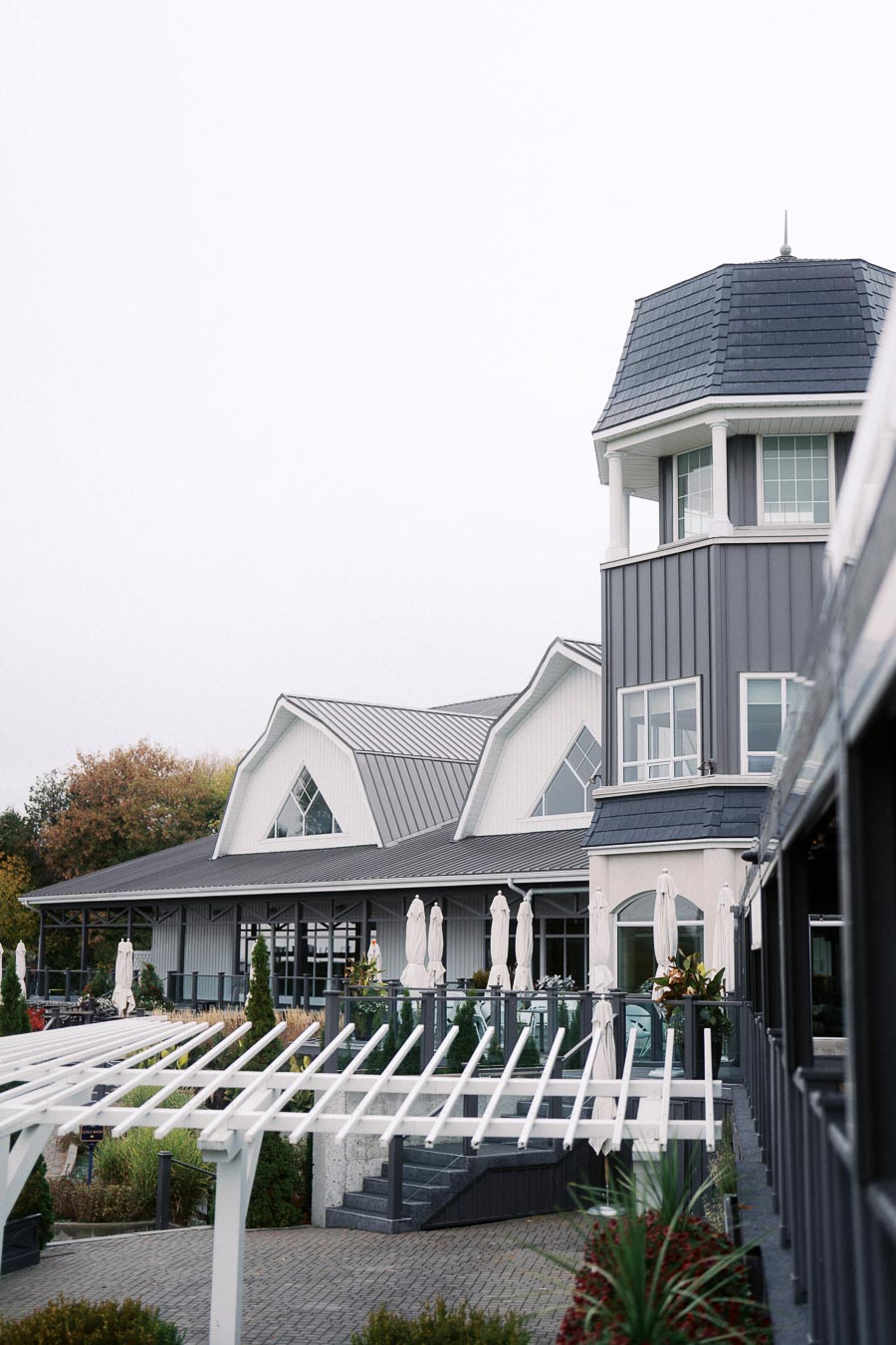 Modern coastal building featuring large windows and a tower structure, surrounded by a landscaped patio with white pergola and outdoor seating, under an overcast sky.