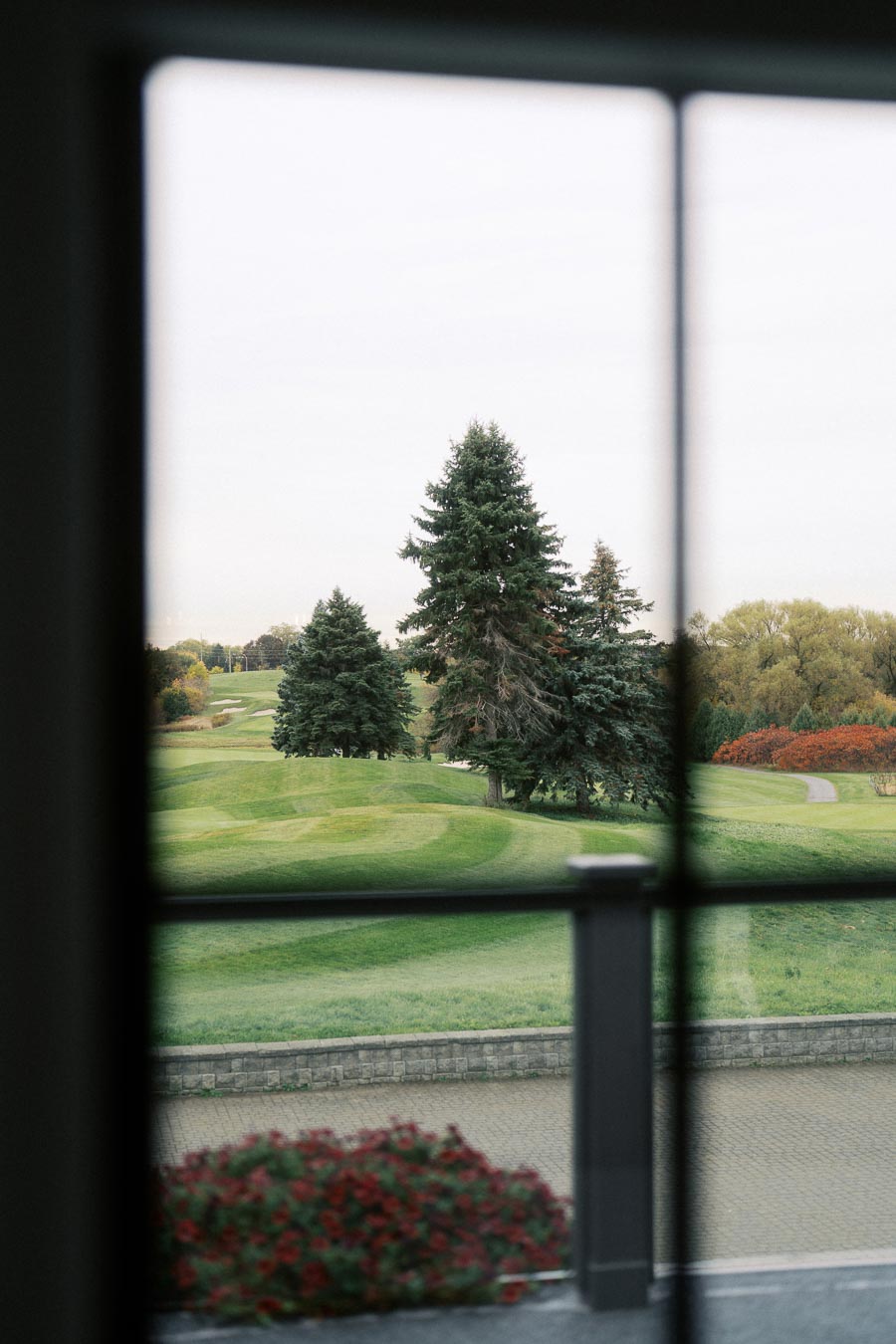 View of a scenic golf course through a window, featuring lush green fairways, tall pine trees, and vibrant autumn foliage.