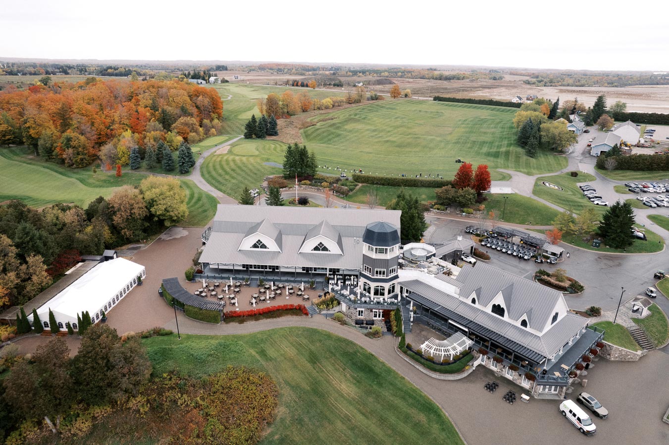 Aerial view of a large, elegant golf clubhouse surrounded by lush green fairways and autumn-colored trees. The clubhouse features multiple wings with decorative roofing and a main tower, with outdoor seating areas visible. Nearby, a parking lot and well-maintained lawns are situated in a picturesque countryside setting.