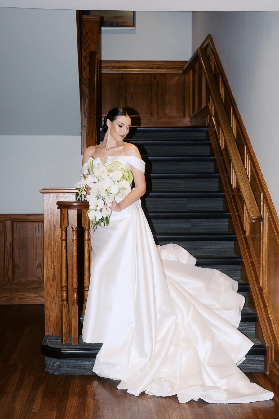 A bride in an elegant white off-the-shoulder wedding gown stands gracefully on a wooden staircase, holding a large bouquet of white flowers. The long train of the dress cascades down the steps, creating a sophisticated and timeless bridal portrait.