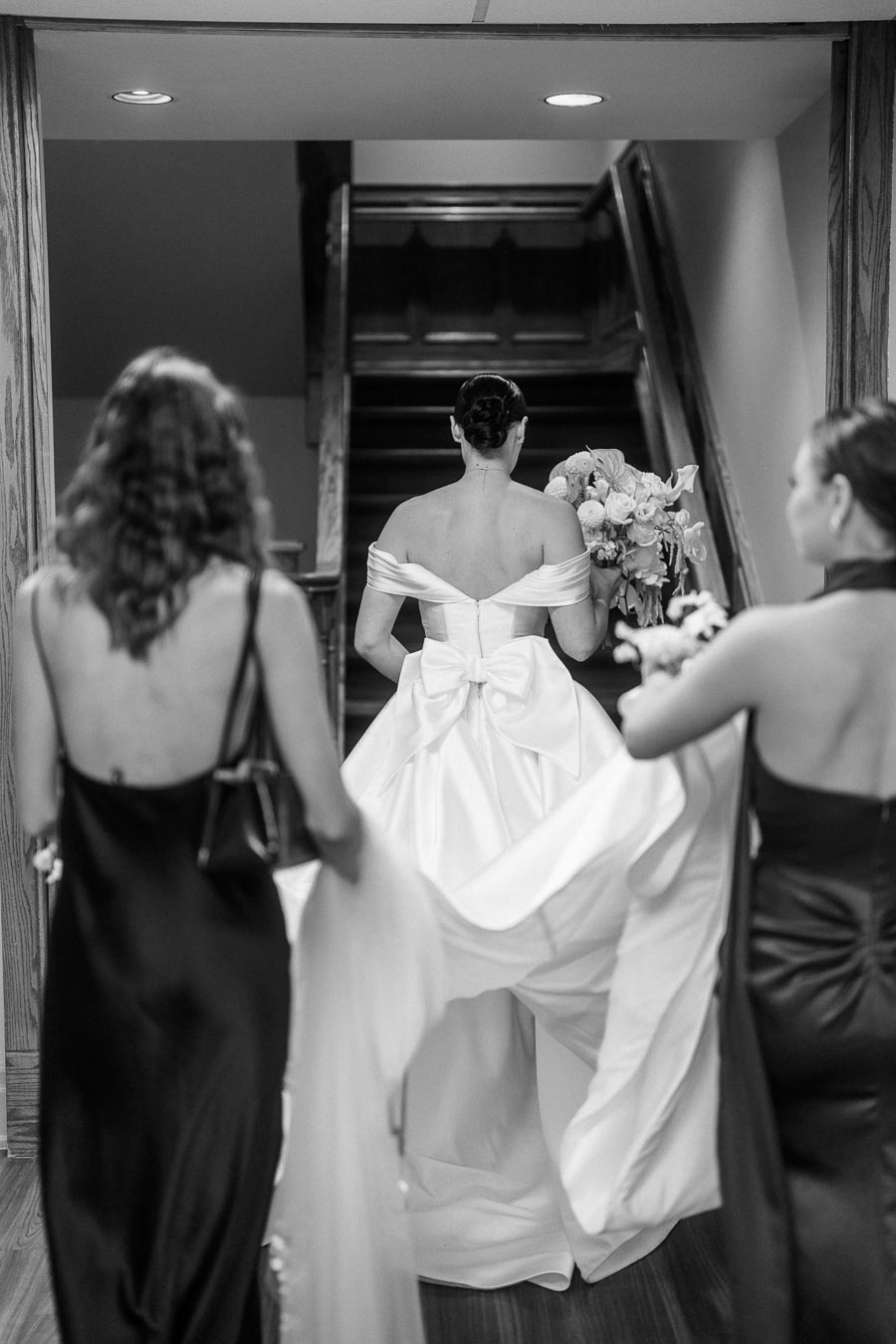 Bride in elegant white wedding dress with large bow, holding bouquet, escorted by two bridesmaids in dark gowns, ascending stairs at indoor venue.