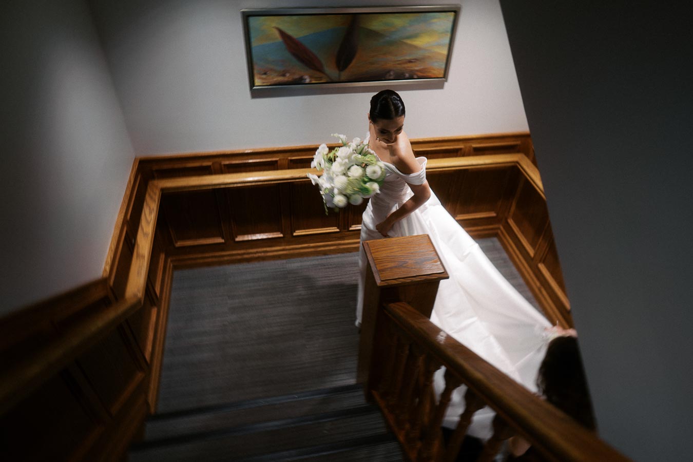 Bride in elegant white wedding dress holding a bouquet while descending wooden staircase indoors, capturing a moment of wedding day preparation.