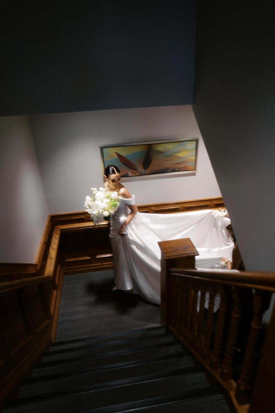 Bride descending staircase in elegant white gown, holding a bouquet of white flowers, with wooden railing and modern artwork on the wall.