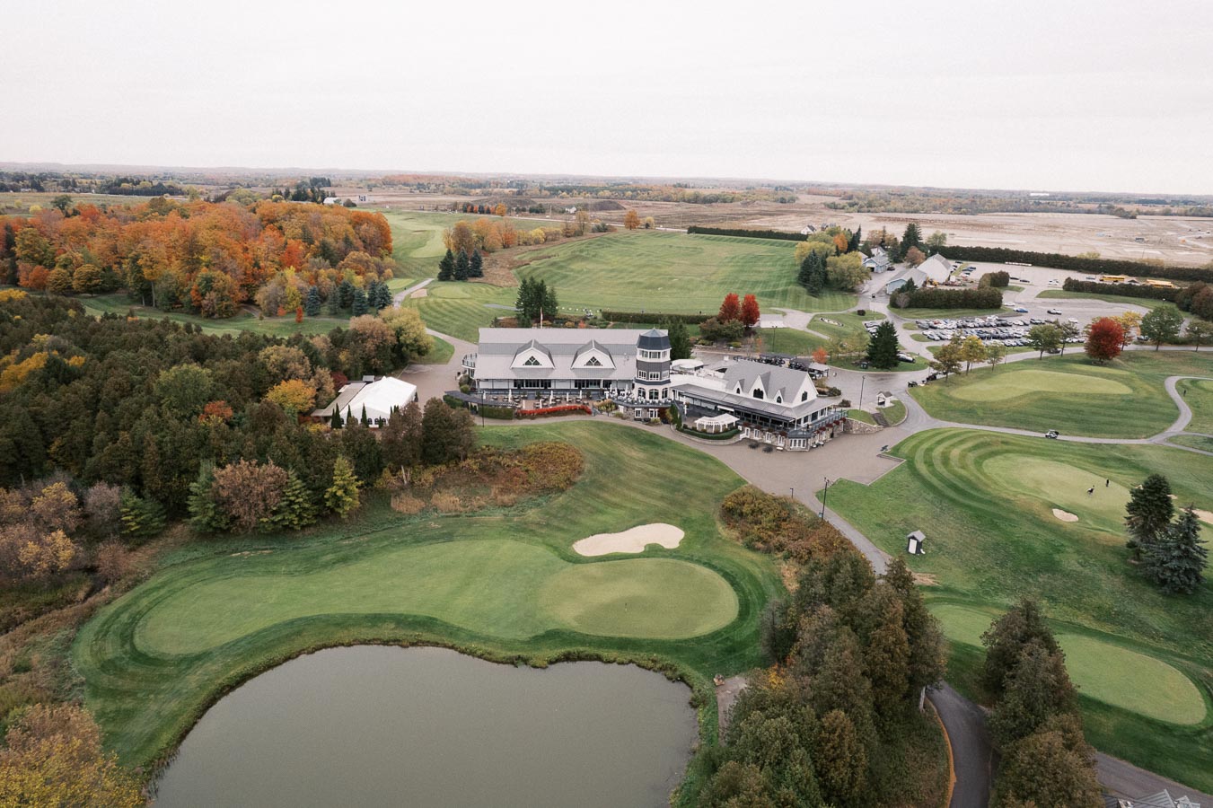 Aerial view of a scenic golf course with lush green fairways, surrounded by autumn-colored trees, featuring a large clubhouse and a tranquil pond, emphasizing the beauty of a serene golfing destination.