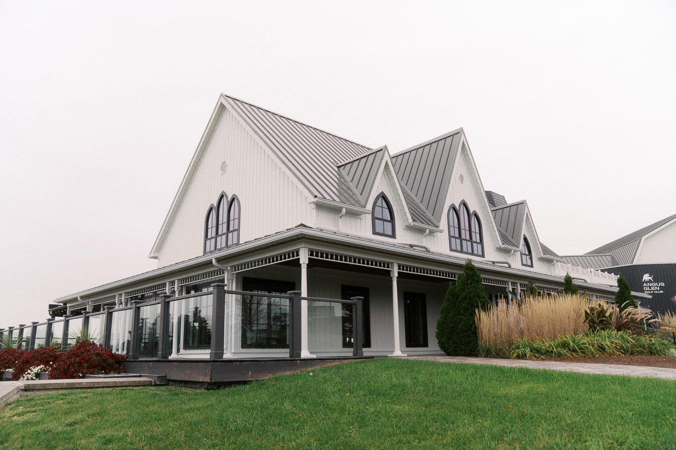 Elegant white building with Gothic-style windows and a metal roof, surrounded by a well-manicured lawn and landscaping, representing a clubhouse or event space setting on a cloudy day.