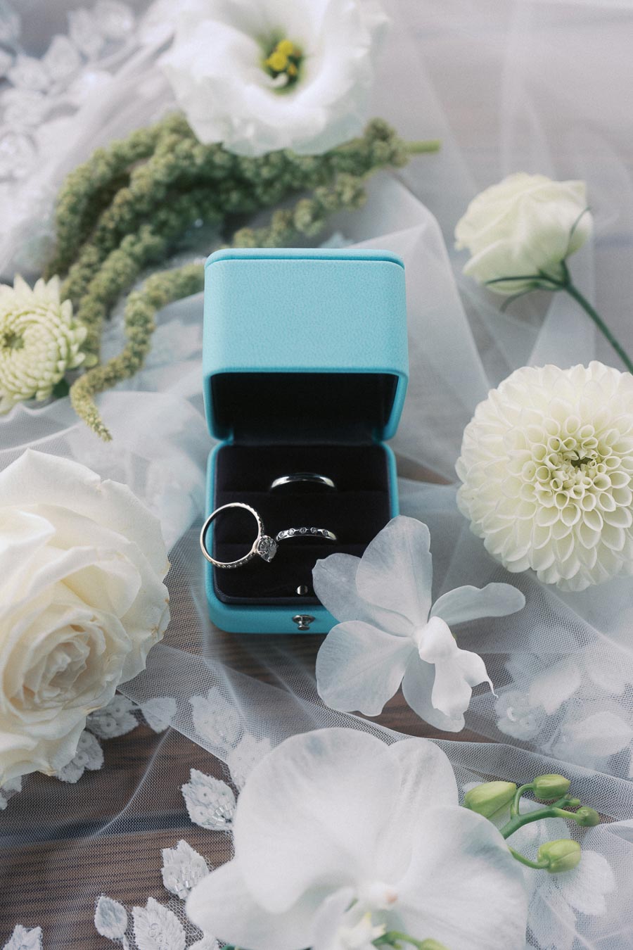Elegant diamond engagement ring in a blue box surrounded by white roses and delicate flowers, set on sheer lace fabric.