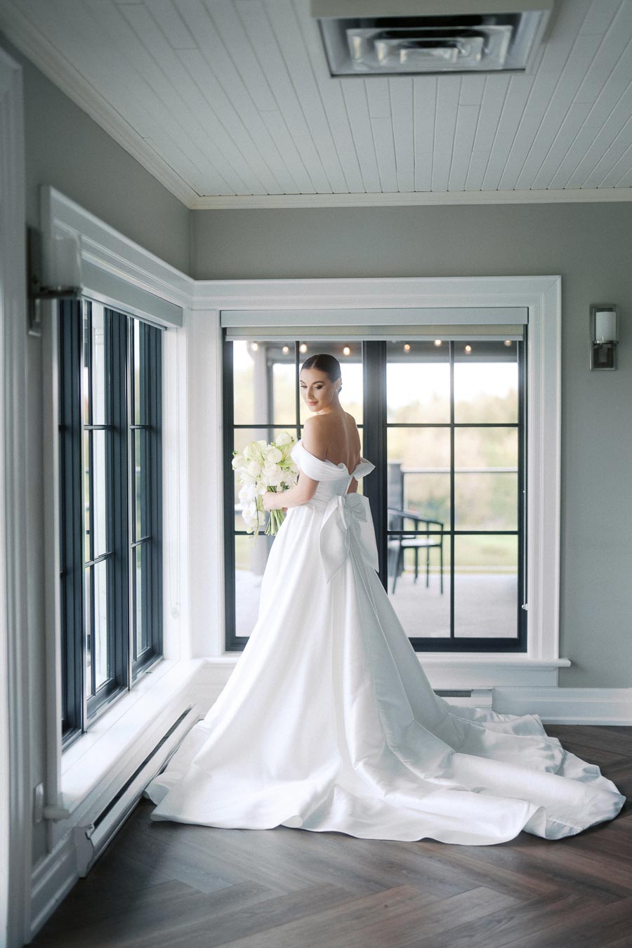 Bride in elegant white off-the-shoulder gown holding a bouquet, standing in a bright room with large windows and wooden floors.