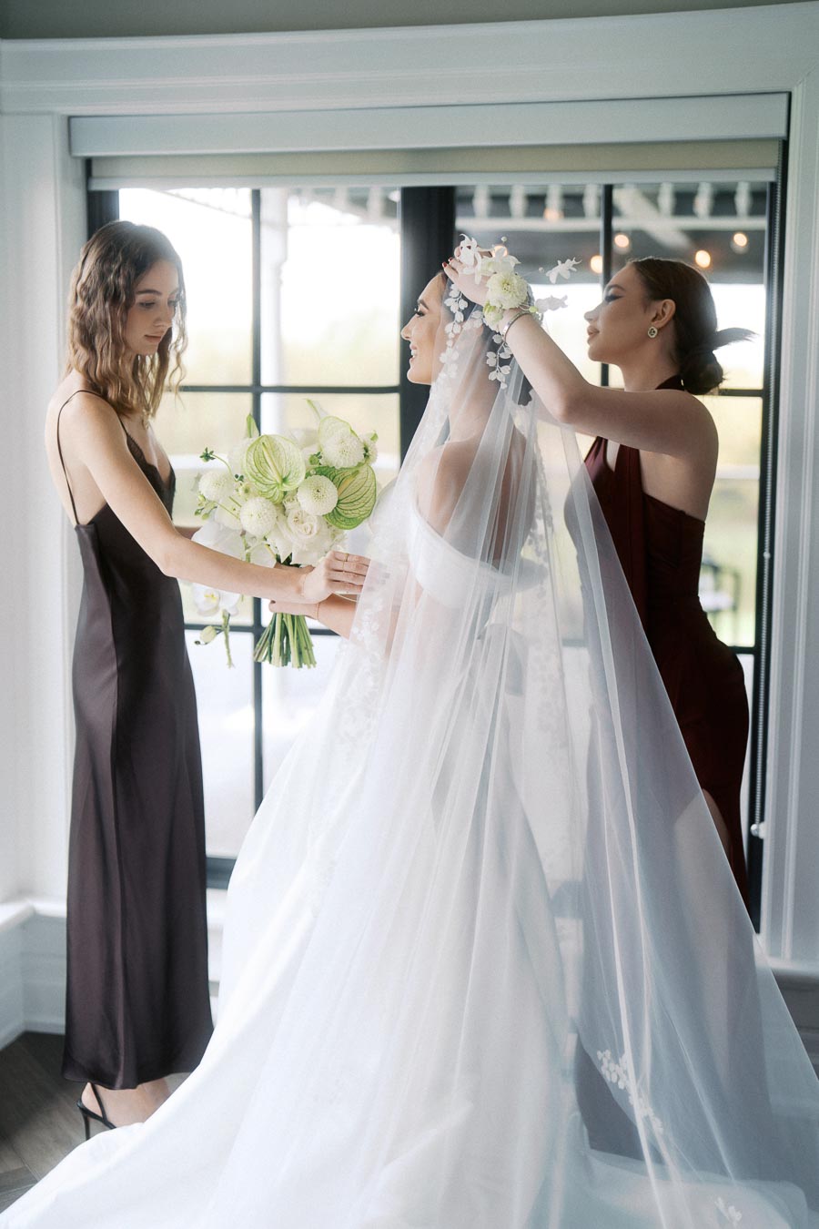 Bride getting ready with bridesmaids in an elegant dress, holding a bouquet of white flowers, and wearing a delicate veil near a large window in a bright room.
