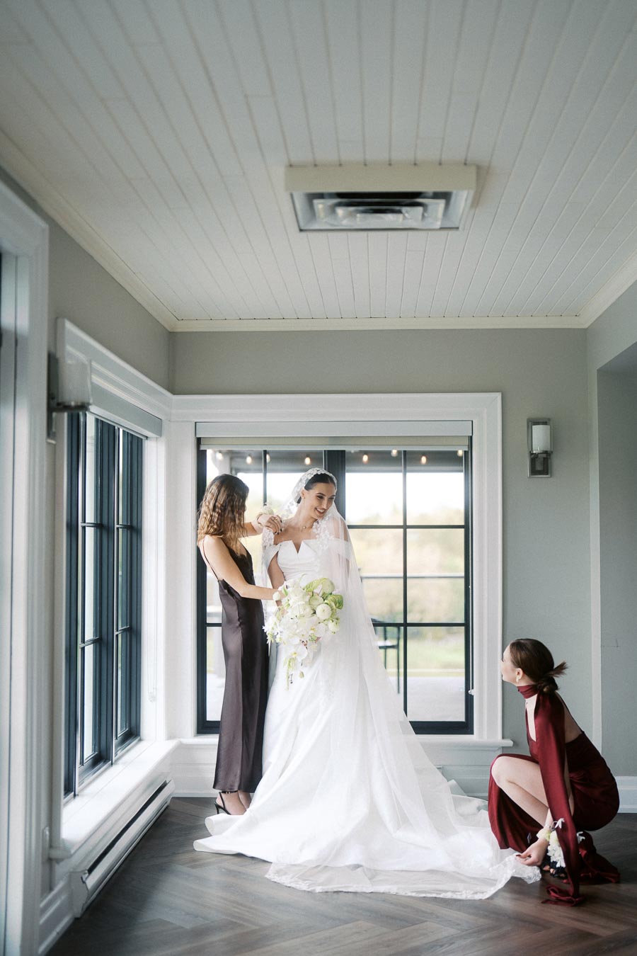 Bride smiling in a white wedding dress holding a bouquet, with two bridesmaids adjusting her dress in a brightly lit room with large windows and wooden flooring.