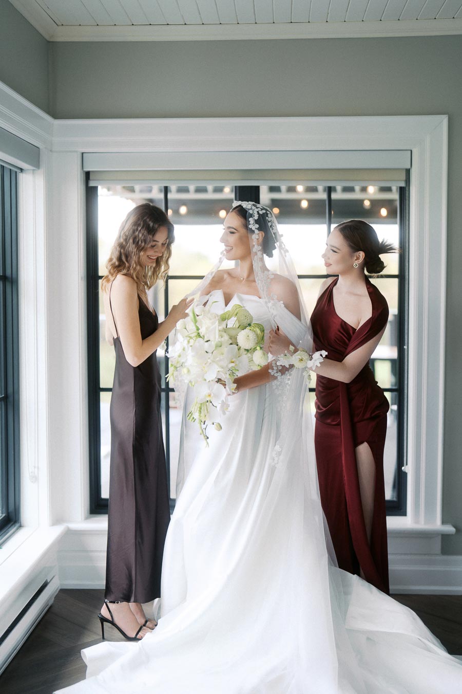 Bride in elegant white gown with veil holding a bouquet, standing between two bridesmaids in rich-colored dresses inside a stylish room with large windows.
