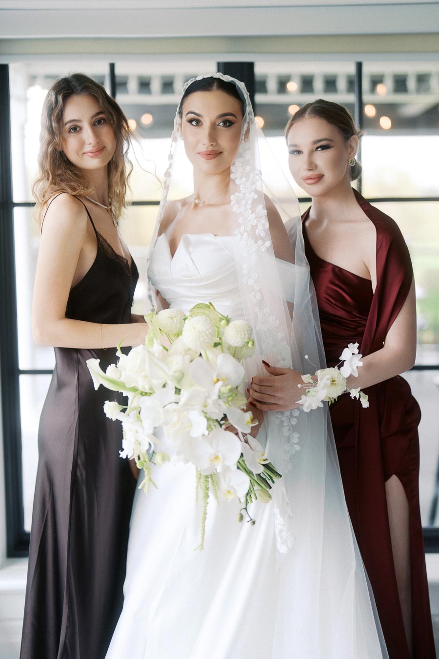 Three women pose together, with the bride in a white wedding dress and floral veil holding a bouquet of white flowers, flanked by two bridesmaids in elegant evening gowns, one in brown and the other in burgundy, inside a bright room with large windows.
