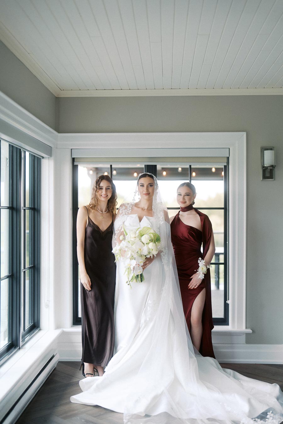 Bride in elegant white gown holding a bouquet, flanked by two bridesmaids in stylish dresses, standing in a bright room with a large window backdrop.