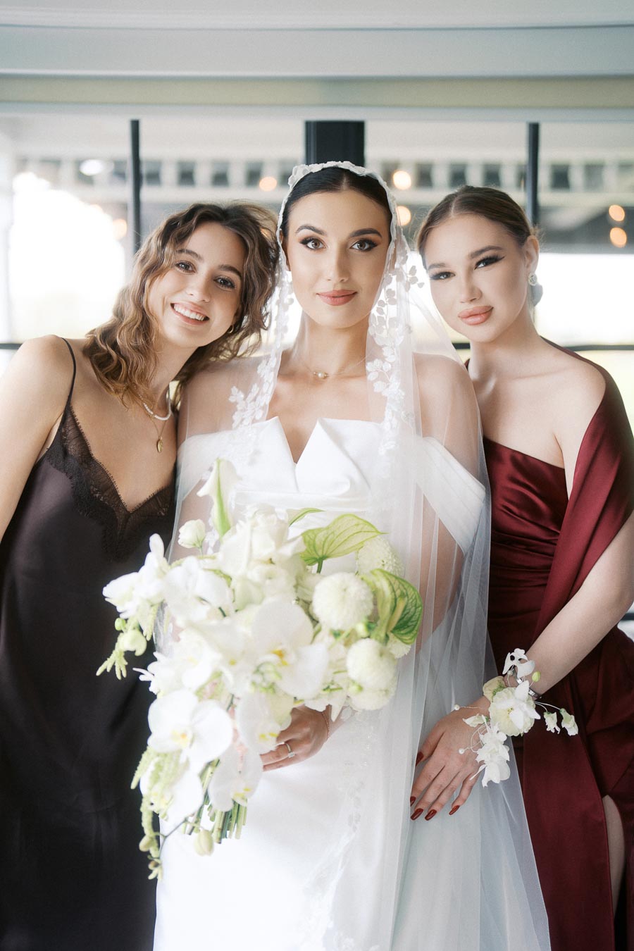 A bride in an elegant white wedding dress and veil holding a bouquet of white flowers, stands smiling with two bridesmaids; one in a black dress and the other in a burgundy outfit, in a bright and modern venue.