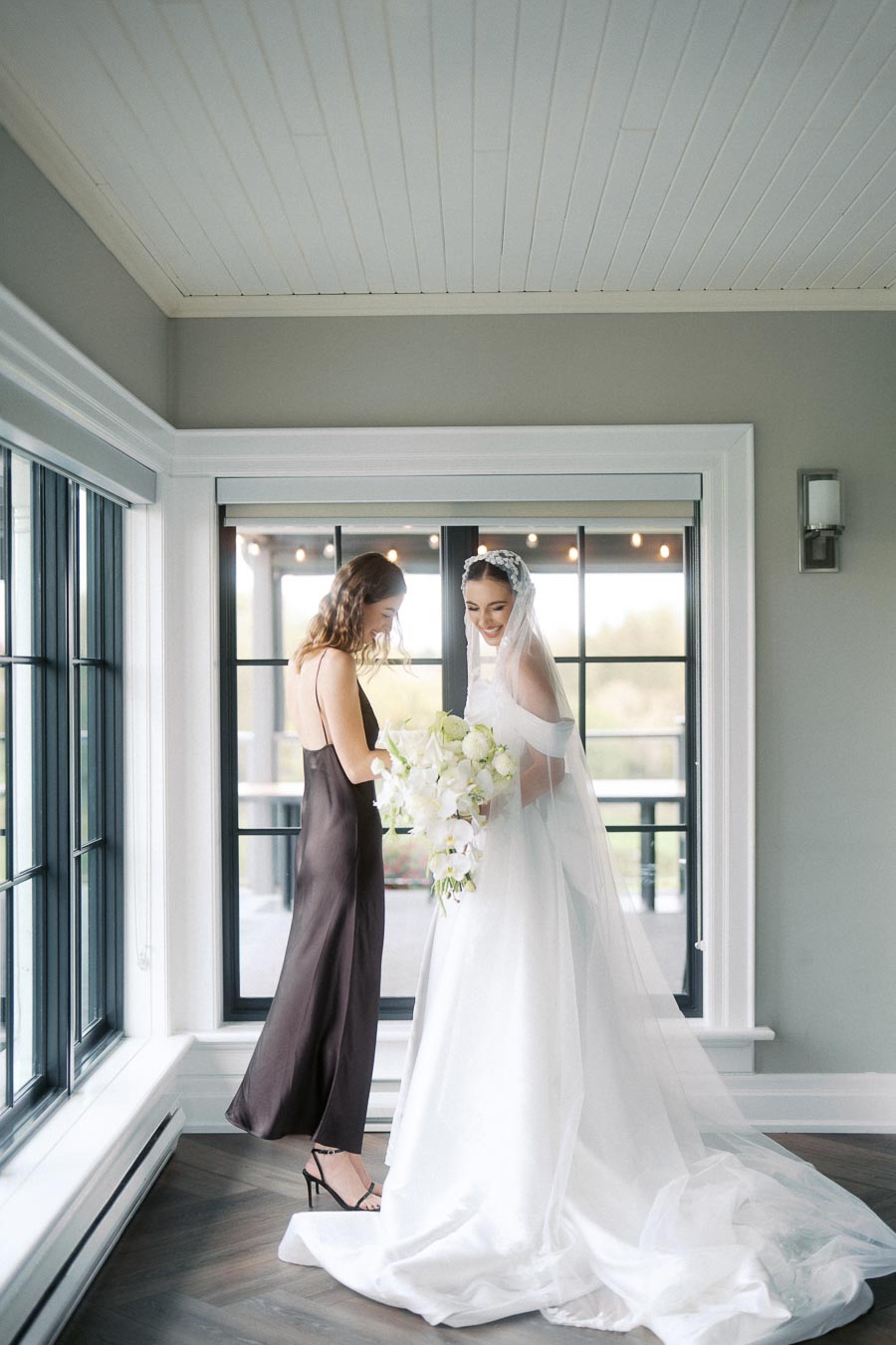 A bride in a beautiful white gown and veil stands next to a woman in a dark sleeveless dress, both smiling and holding a bouquet of white flowers, in a bright room with large windows.