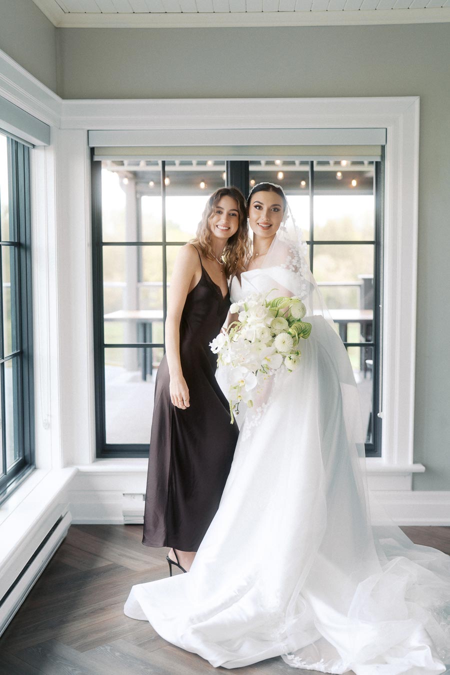 Bride in elegant white wedding gown holding bouquet, standing next to a woman in a stylish dark dress inside a sunlit room with large windows.