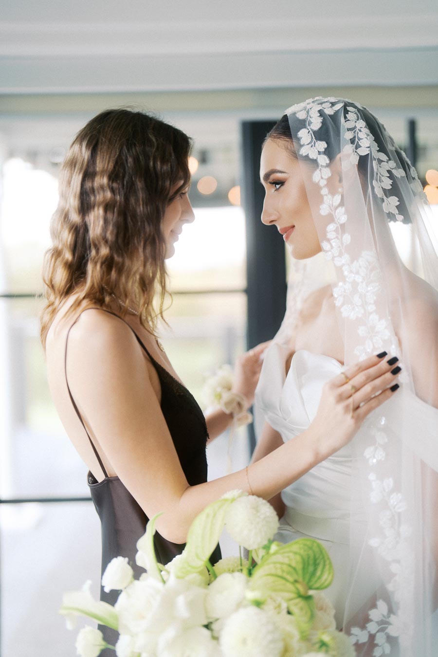 Bride in an elegant white dress and veil sharing a tender moment with a bridesmaid in a black dress, holding a bouquet of white flowers.