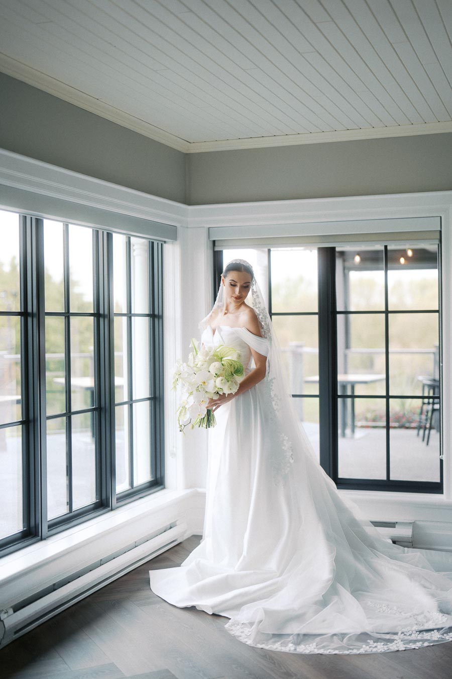 Bride in an elegant white wedding gown holding a bouquet, standing in a bright room with large windows and natural light streaming in.