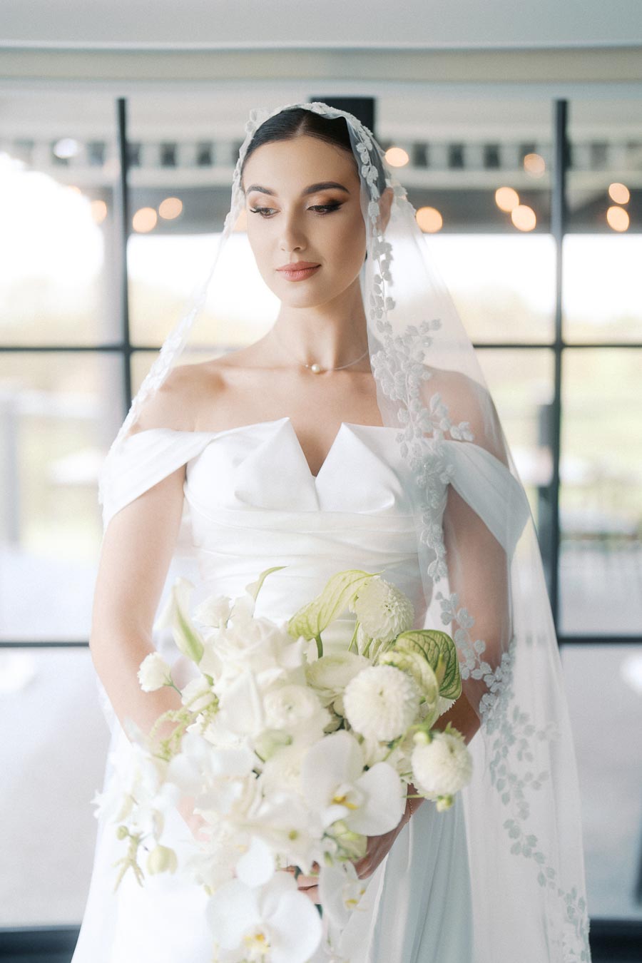 Elegant bride in a white wedding dress with delicate lace veil, holding a bouquet of white flowers in a softly lit indoor setting.