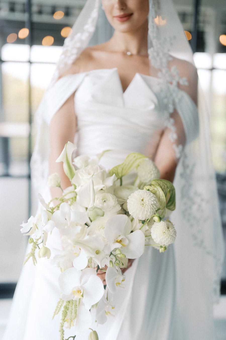 Bride in elegant white wedding dress holding a bouquet of white flowers, featuring orchids and roses, with delicate lace veil in a softly lit setting.
