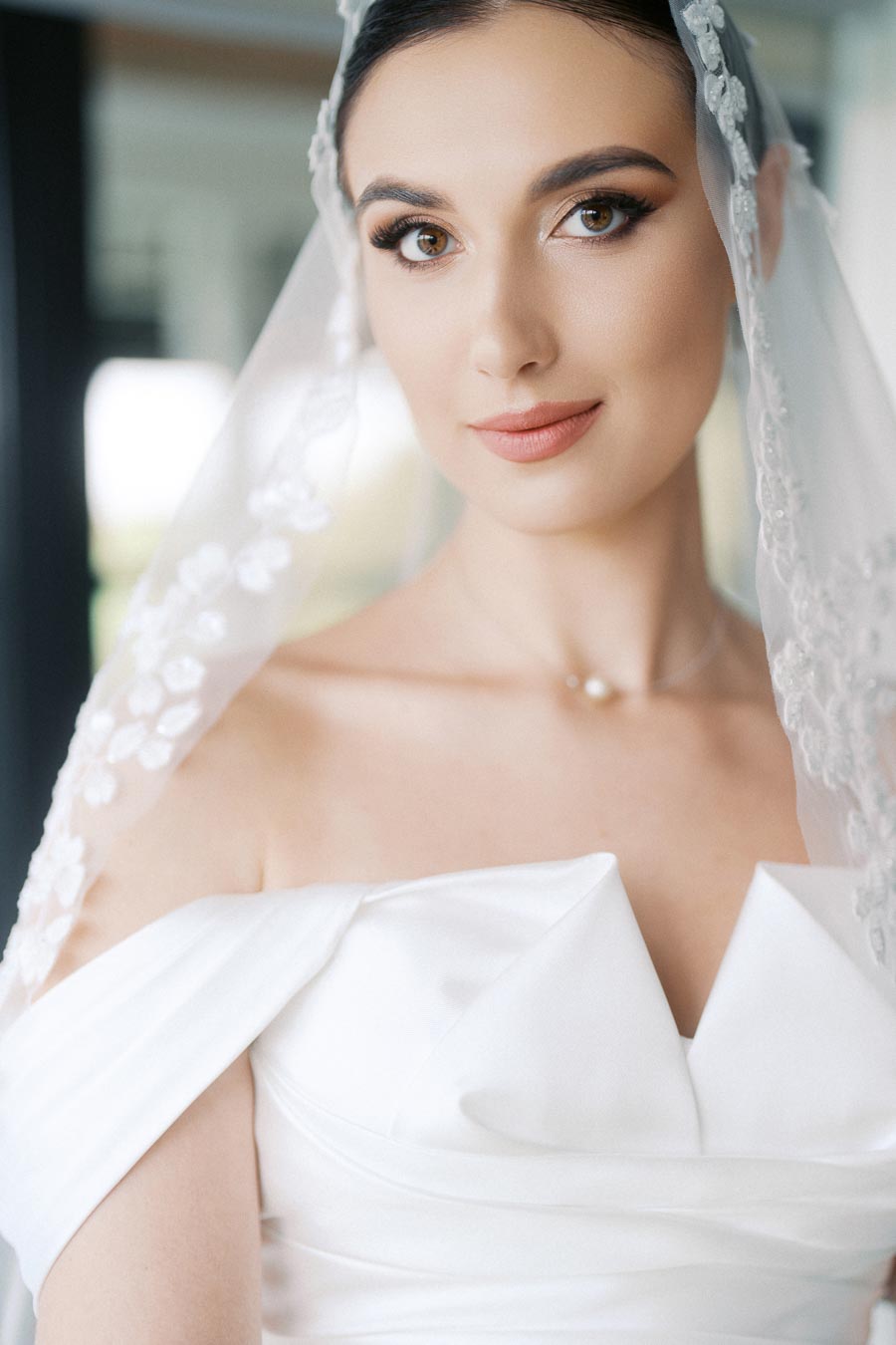 Elegant bride wearing a white wedding dress and lace veil, showcasing flawless makeup and a gentle smile.