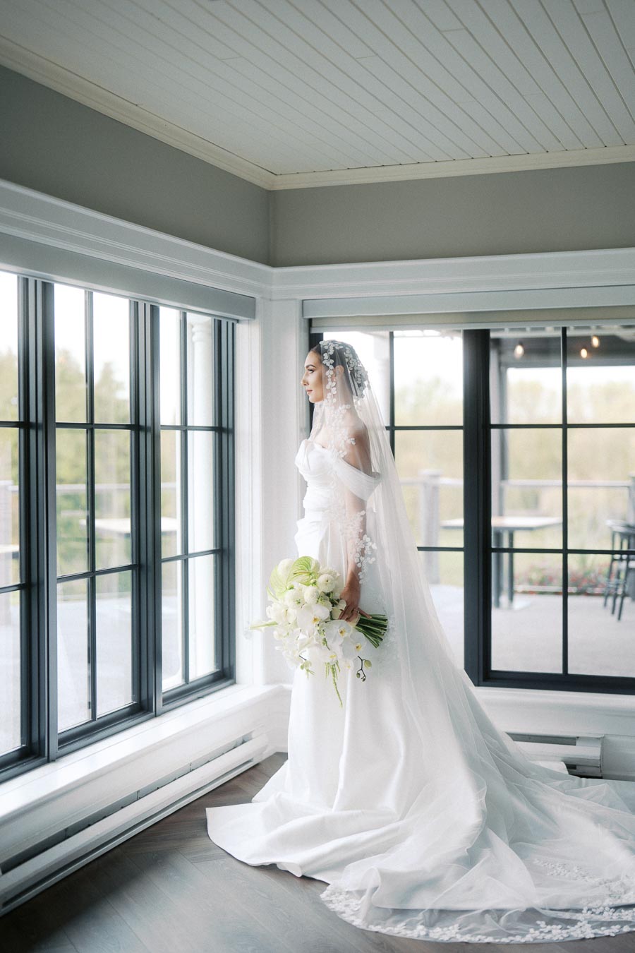Elegant bride in white gown holding a bouquet, standing by large windows and gazing outside; wedding photography with natural lighting.