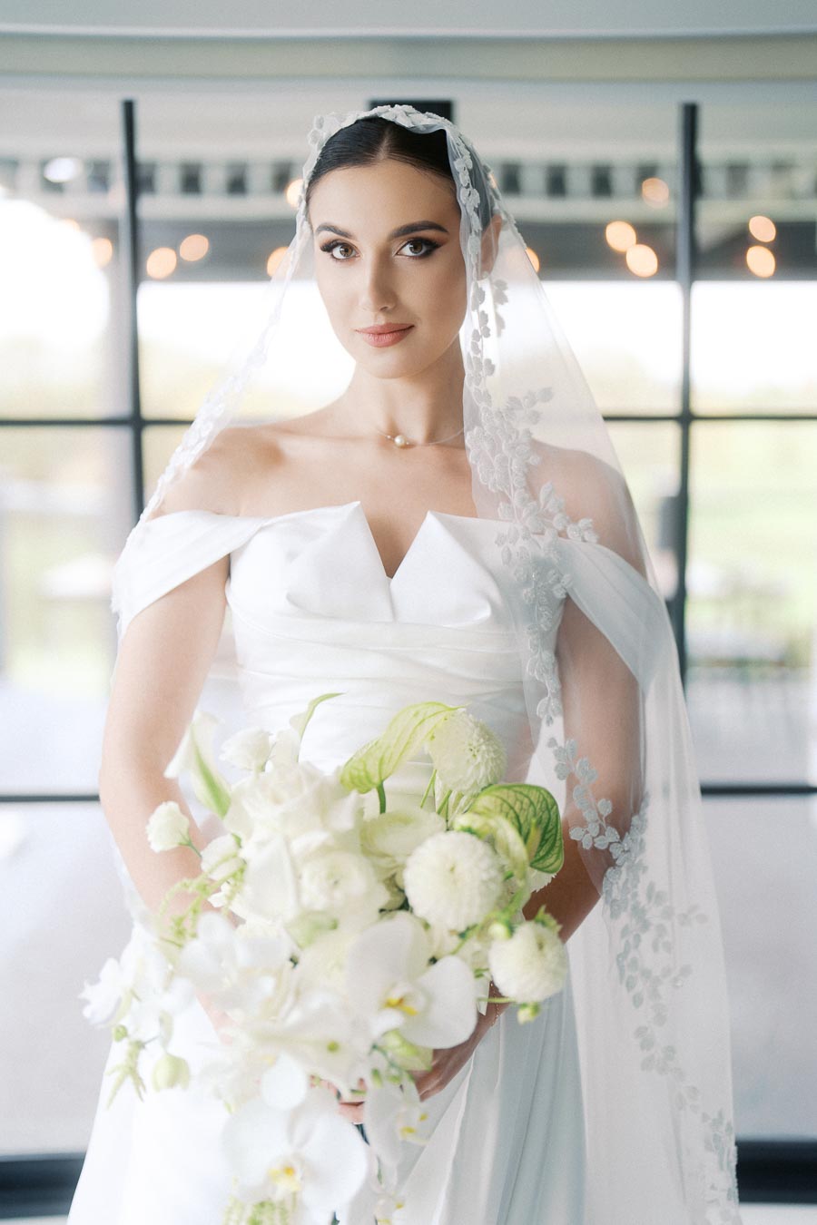 Bride in a white off-shoulder wedding dress with a lace veil, holding a bouquet of white flowers, standing in front of a large window with soft lighting.