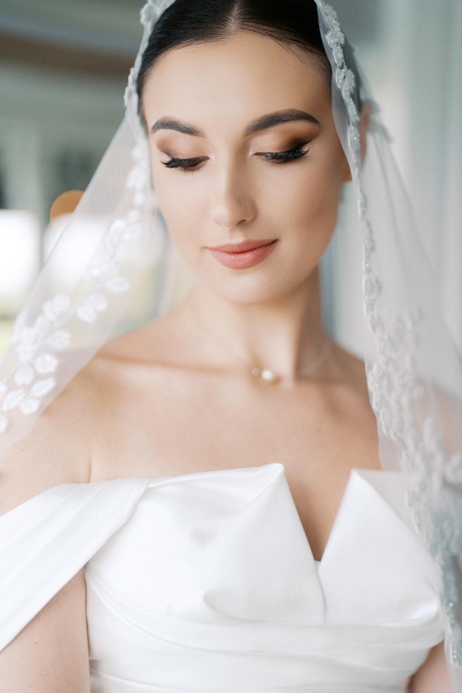Elegant bride in a white wedding gown and delicate lace veil gazes downward with soft makeup and serene expression, capturing a timeless bridal moment.
