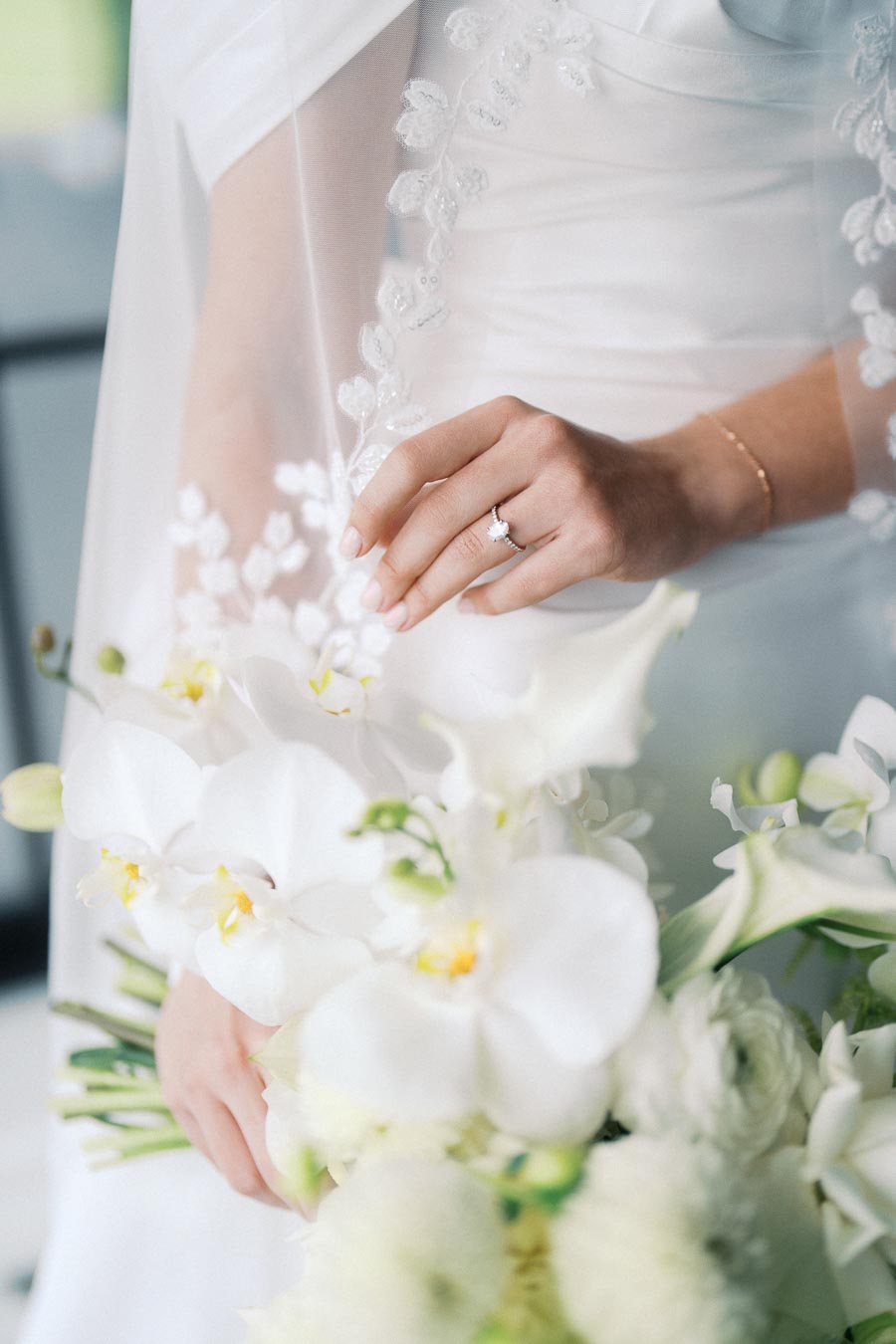 Bride's hand with wedding ring gently touching a white orchid bouquet, showcasing delicate lace details on the wedding dress veil.