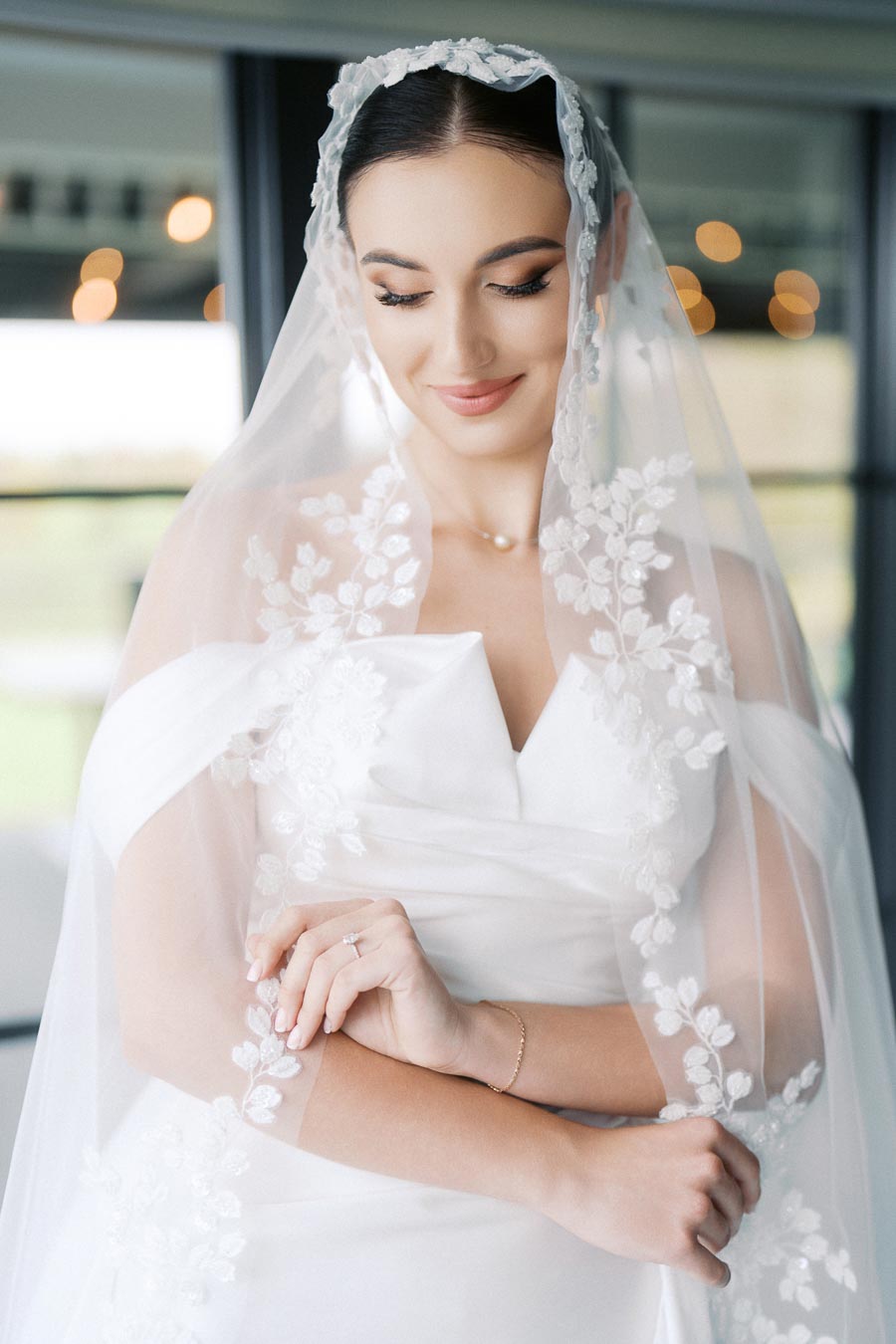 Bride in an elegant white wedding dress and lace veil, smiling softly with folded arms, showcasing a delicate pearl necklace and a delicate ring, in a softly lit setting.