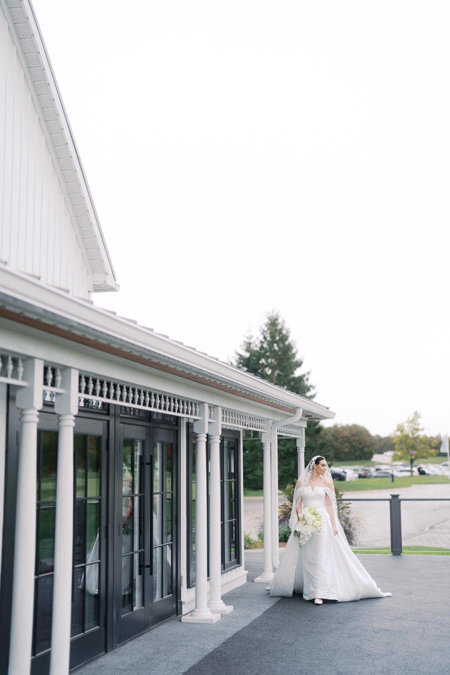 Bride in elegant white dress and veil walking along a veranda with white columns at an outdoor wedding venue.